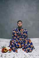 Free photo contemplated young man sitting on floor with crossed legs near the gerbera flowers