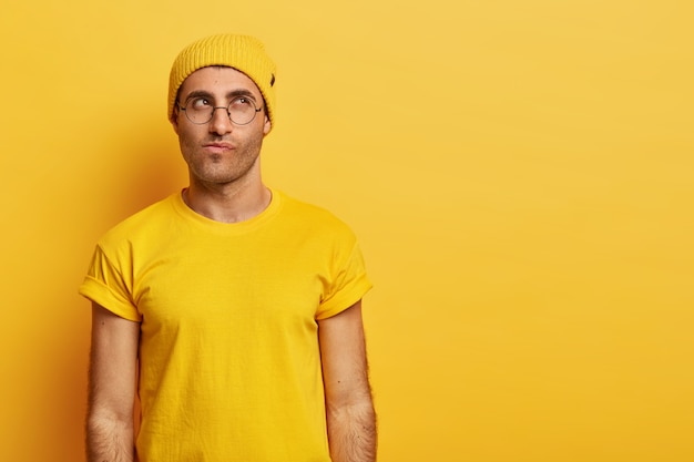 Free photo contemplative caucasian guy looks with thoughtful expression upwards, focused above, purses lips, tries to make decision