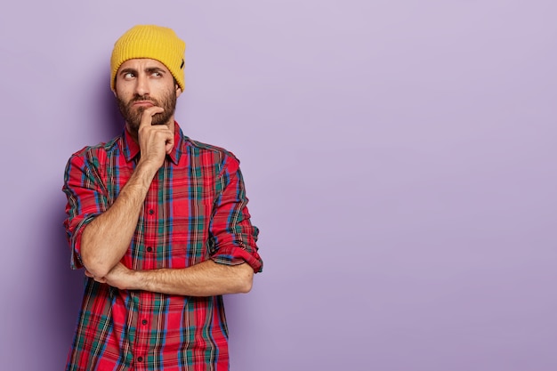 contemplative young man holds chin, looks aside with thoughtful expression, thinks something over, wears yellow hat and plaid shirt