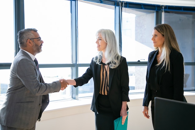 Content businesspeople greeting, meeting and smiling. Successful Indian CEO in eyeglasses handshaking with grey-haired businesswoman, talking and looking at her. Business and partnership concept