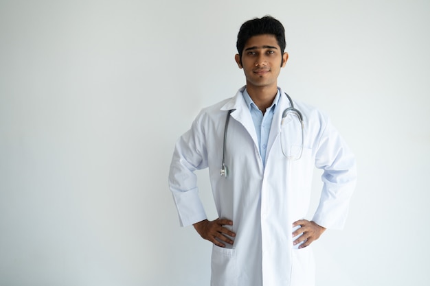 Content successful general practitioner in lab coat holding hands on hips 