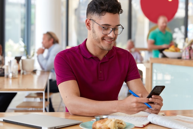 Content young male blogger holds cell phone, downloads files, sends text messages, uses free internet connection, poses against cafe interior, researches information. Technology and work concept