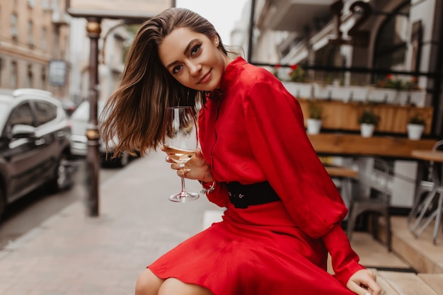 Free photo contented, sweet girl smiles gently. red dress adds brightness for outfit of lady posing with glass of wine