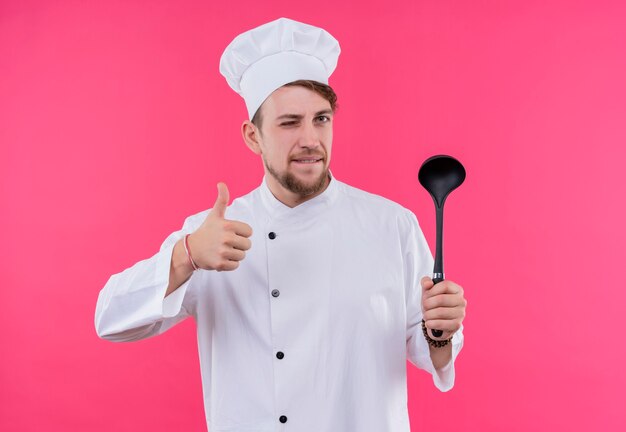 Free photo cook looking at camera shutting one eye showing like gesture with spoon on hand standing over pink wall