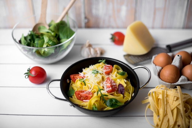 Cooking pot filled with healthy tagliatelle pasta and ingredients on white wooden table