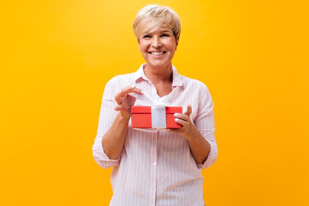 Cool adult lady smiles and holds gift box on orange background