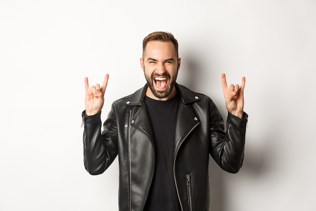 Cool adult man in black leather jacket, showing rock on gesture and tongue, enjoying music festival, standing  