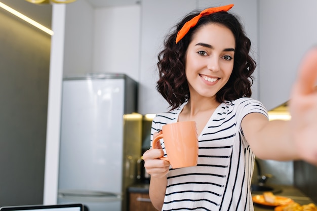 Cool brightful selfie portrait of attractive young woman with cut curly brunette hair smiling with cup of tea in kitchen of modern apartment. Having fun, true positive emotions