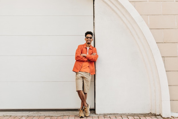 Free Photo cool brunet bearded man in sunglasses beige shorts and orange jacket leans on white wall outside and crosses arms