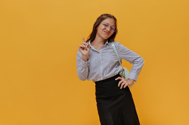 Cool emotional young girl points up on isolated orange background Charming teenager in school uniform looks into camera