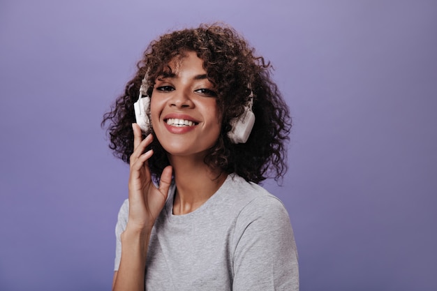 Cool girl in gray top smiles and listens to music on purple wall