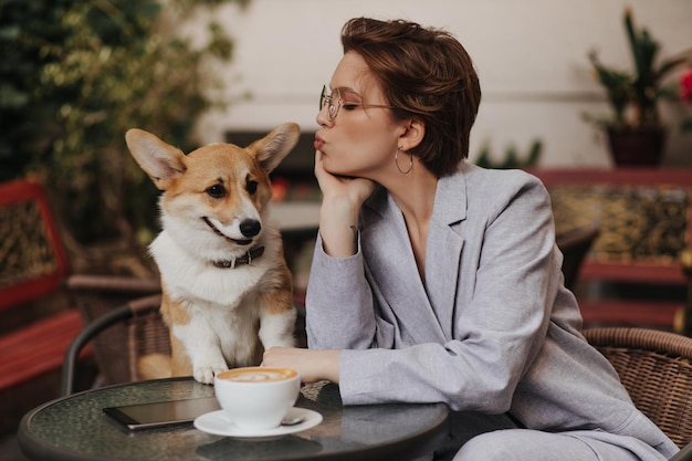 Free photo cool girl in grey suit kisses her dog and chills in street cafe pretty darkhaired woman in jacket resting with corgi outside