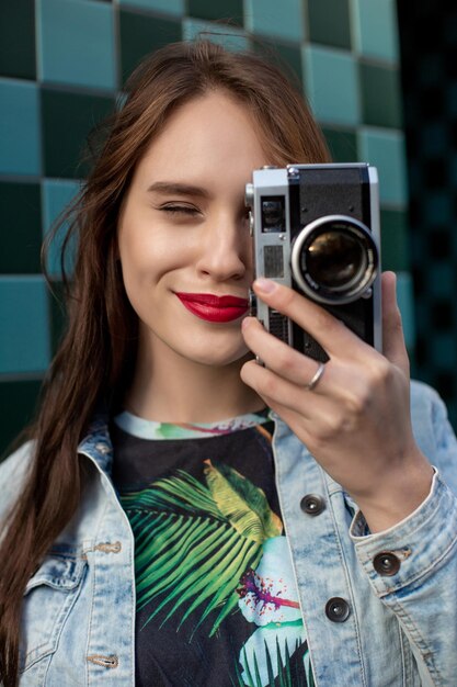 Cool girl model with retro film camera wearing a denim jacket, dark hair outdoors over city wall in a cage background. Having fun in the city with camera, travel photo of photographer.