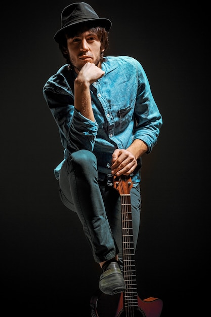 Free photo cool guy with hat standing with guitar on dark studio background