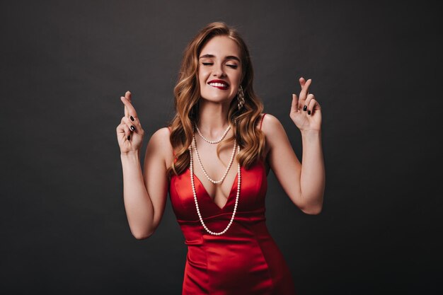 Cool lady in red silk outfit crosses fingers on black background Blonde curly woman in satin dress happily posing on isolated backdrop