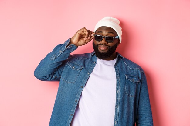 Cool and sassy african-american bearded man, looking confident, touching sunglasses and stare at camera please, standing over pink background