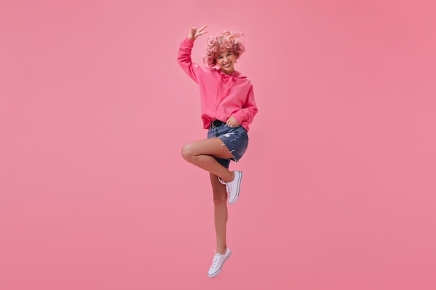 Free photo cool shorthaired girl in pink hoodie and denim shorts jumping and smiling happy woman with pink hair shows peace sign on isolated