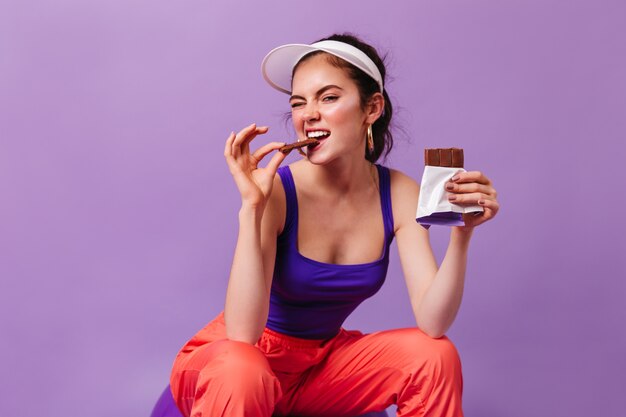 Cool woman in sports outfit in style of 80s bites off delicious milk chocolate bar sitting on purple wall