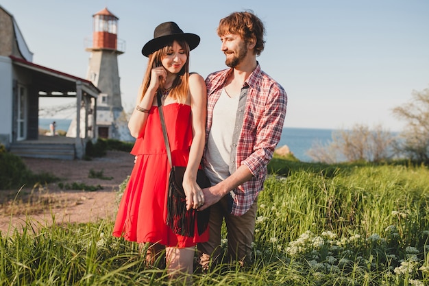 Cool young stylish couple in love in countryside, indie hipster bohemian style, weekend vacation, summer outfit, red dress, green grass, holding hands, smiling