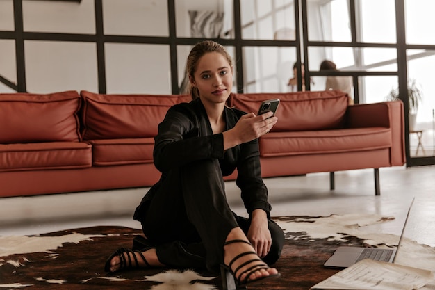 Cool young woman in stylish black silk suit sits on floor on carpet in living room holds phone an