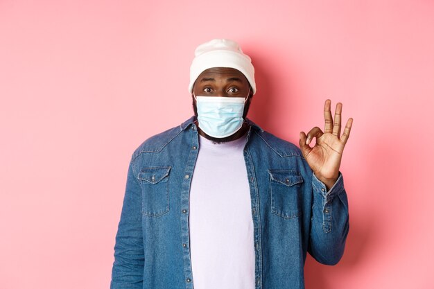 Coronavirus, lifestyle and social distancing concept. Impressed african-american man in face mask, showing okay sign, like and agree, standing over pink background