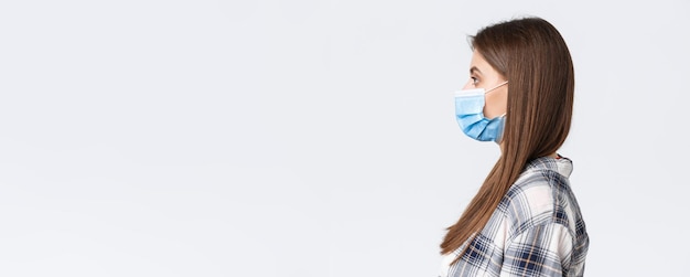 Coronavirus outbreak leisure on quarantine social distancing and emotions concept Profile of seriouslooking young pretty woman in medical mask standing in line white background