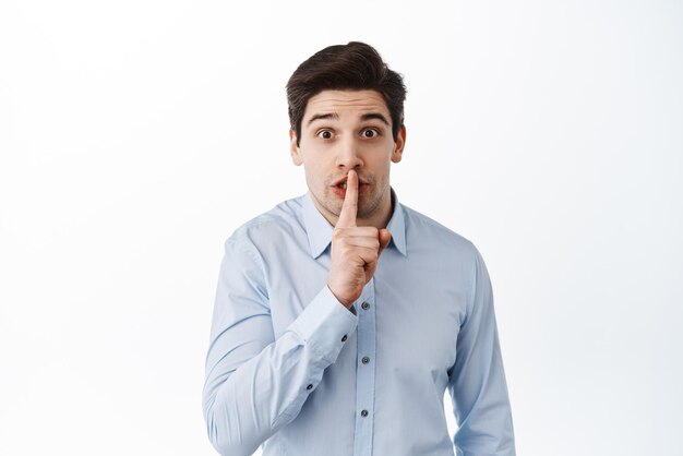 Corporate man in shirt asking to keep quiet gossiping in office shushing holding finger to lips and looking excited white background