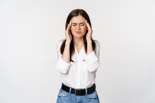 Corporate woman grimacing, touching head, feeling headache, severe migraine, standing over white background