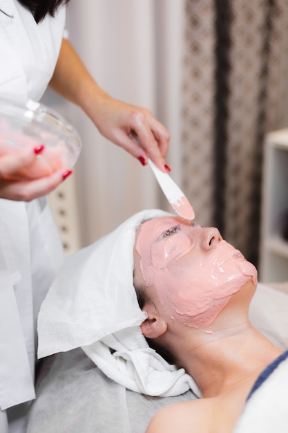 Cosmetologist applies alginate mask with spatula on face of woman.