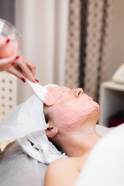 Cosmetologist applies alginate mask with spatula on face of woman.