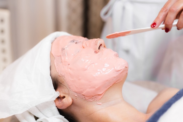 Cosmetologist applies alginate mask with spatula on face of woman.