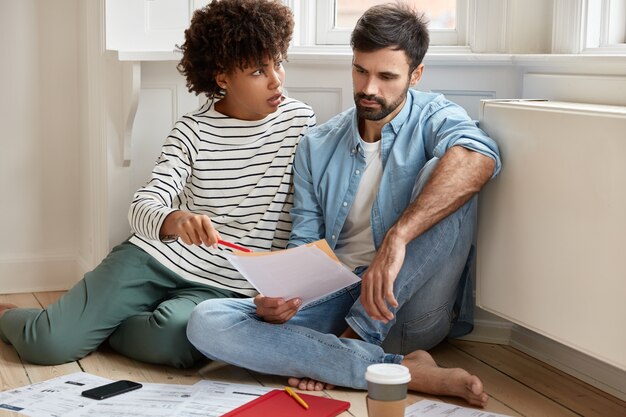 Couple calculate their bills at home, look desperately at papers, have much unpaid taxes, check documentation, pose on floor, drink take out coffee from disposable cup, think about repairing flat