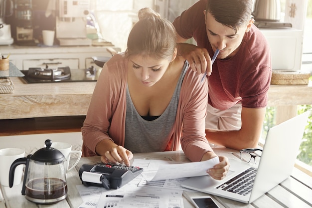 Couple facing financial stress. Young female dressed casually planning family budget in kitchen, using calculator. Her husband holding pen and standing next to her