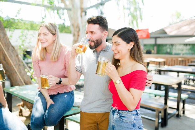 Couple in love hugging and laughing while drinking beer with some friends at an outdoor bar. Boyfriend and girlfriend hanging out with a group of friends