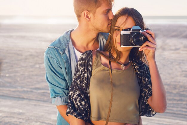 Couple in love posing on the  evening beach, Young hipster girl and her handsome boyfriend taking photos with retro film camera. Sunset warm light.