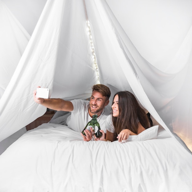 Free photo couple lying on bed toasting alcohol bottles taking selfie on cellphone