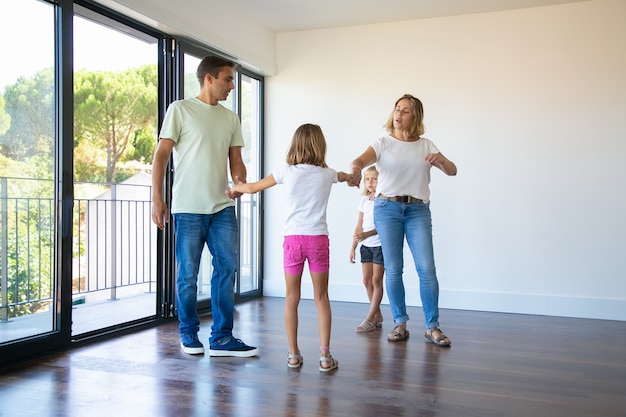 Couple of parents and two kids enjoying their new home, standing in empty room and holding hands, dancing