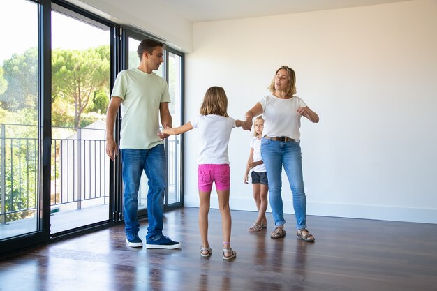 Couple of parents and two kids enjoying their new home, standing in empty room and holding hands, dancing