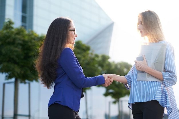 Free photo couple of serious businesswomen greeting one each other with handshake outside of their offices.