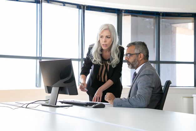 Couple of serious colleagues discussing content on computer monitor, pointing at display and talking while sitting in meeting room with panoramic window. Business communication concept