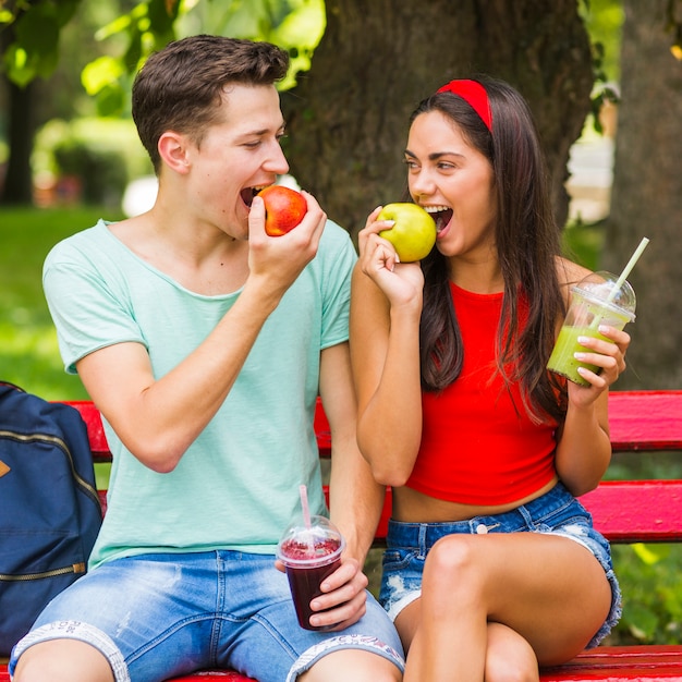 Free photo couple sitting on bench eating ripe apples holding smoothies