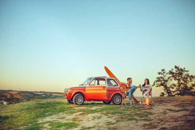 Free photo couple sitting and resting on the beach playing guitar on a summer day near river. love, happy family, vacation, travel, summer concept.