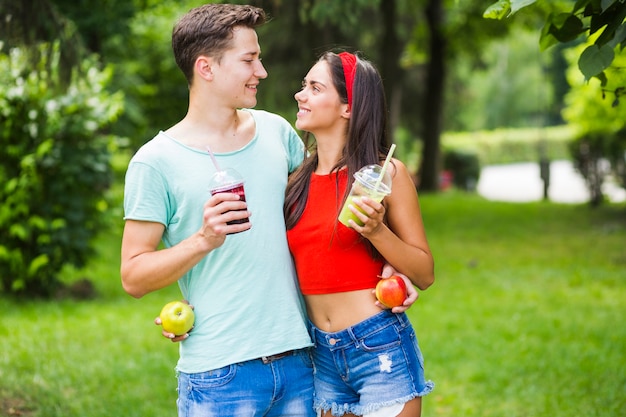 Free photo couple standing in park holding healthy smoothies and apples looking at each other