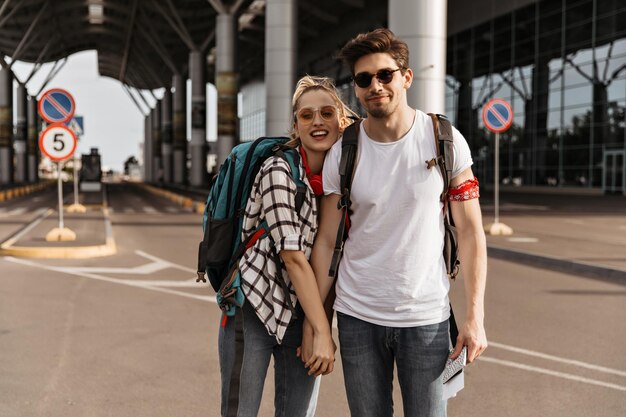 Couple of travelers poses near airport and looks into camera Blonde woman in plaid shirt sunglasses and brunette handsome man in white tee hold hands and poses with backpacks