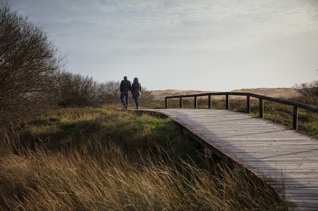 Couple walking on a wooden bridge surrounded by a field and hills under the sunlight