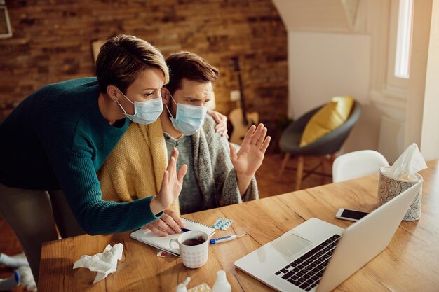 Couple with protective face masks waving during video call over laptop at home