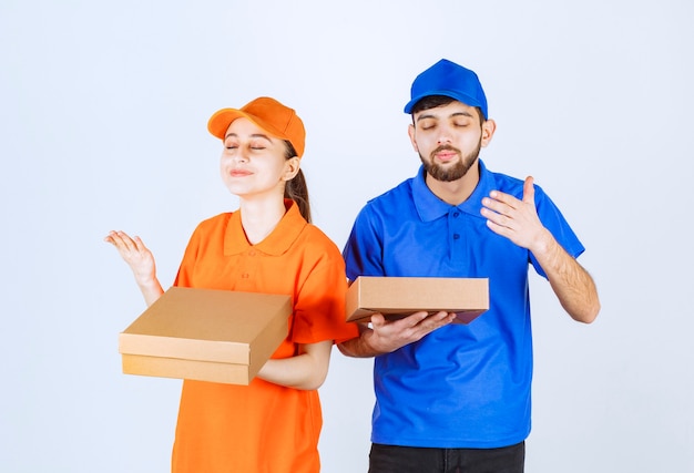 Free photo courier boy and girl in blue and yellow uniforms holding cardboard takeaway boxes and shopping packages and smelling the food.