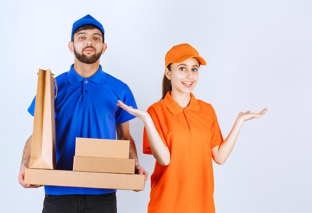 Free photo courier boy and girl in blue and yellow uniforms holding cardboard takeaway boxes and shopping packages.