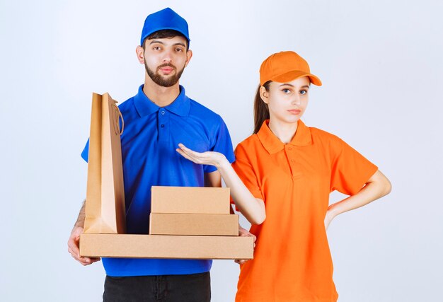 Free photo courier boy and girl in blue and yellow uniforms holding cardboard takeaway boxes and shopping packages.