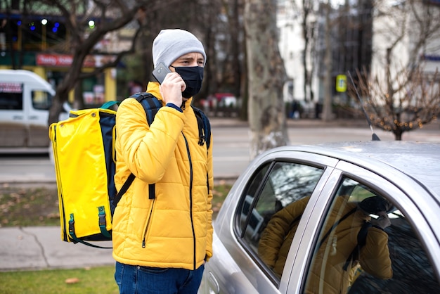 Free photo courier with yellow backpack and black medical mask near a car talking on the phone